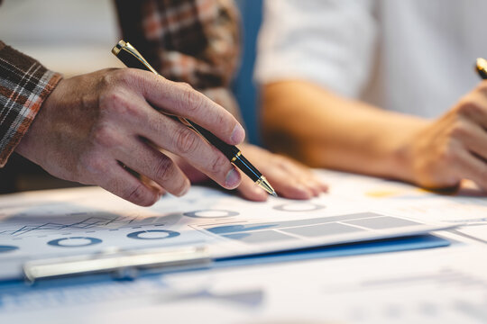 Close-up of two business people reviewing charts and financial documents at a desk, with one person pointing using a pen.