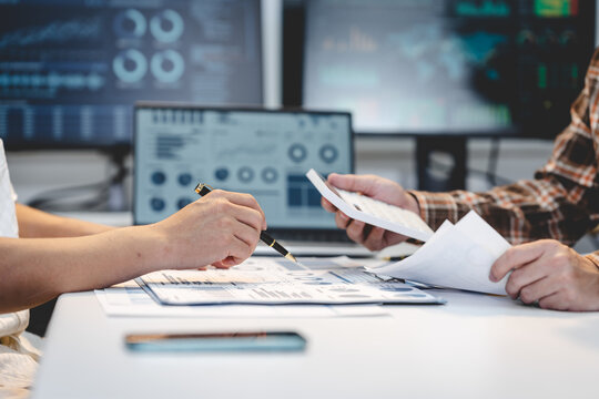 Close-up of coworkers reviewing financial papers and using a calculator at a desk, with computer screens showing data charts in a modern office.