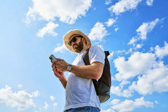 Male Tourist with Backpack Checking Smartphone Outdoors Against Blue Sky