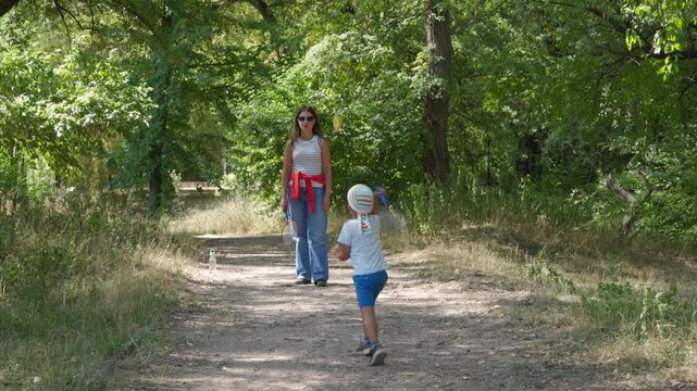 A loving mother watches her playful son run with a badminton racket. They enjoy a happy bonding game on a shady path in a green park.
