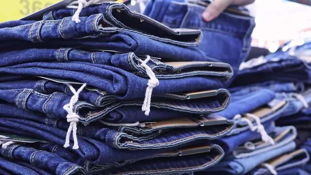 Man with tattooed hand choosing blue denim jeans from folded stacks on display in clothing store. Close-up shopping for casual fashion apparel in retail.