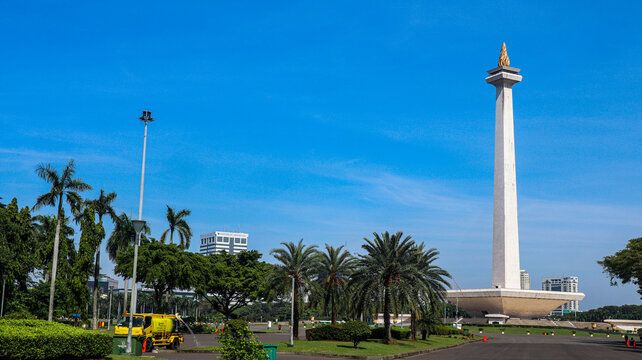 The iconic National Monument (Monas) in Jakarta, Indonesia, is surrounded by a spacious plaza, green landscapes, palm trees, and modern city buildings beneath a bright blue sky. 