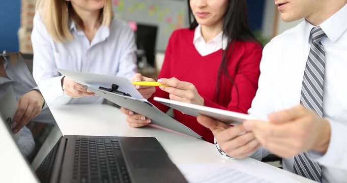 In a vibrant office setting, three professionals in red and white outfits gather around a laptop. They actively share insights, pointing at documents and exchanging ideas.