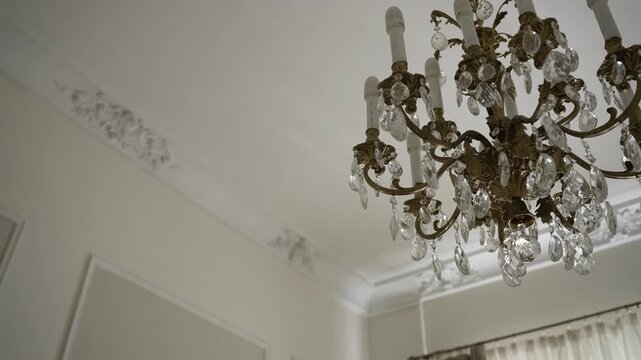 Ornate crystal chandelier moving closer beneath decorative plaster ceiling in elegant interior, showing gilded arms, carved scrollwork, faceted drops, soft reflections, and refined luxury detail