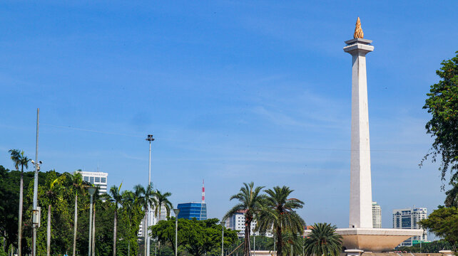 The iconic National Monument (Monas) in Jakarta, Indonesia, is surrounded by a spacious plaza, green landscapes, palm trees, and modern city buildings beneath a bright blue sky. 