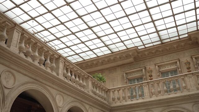 Low-angle pan across ornate neoclassical atrium with frosted skylight grid and stone balustrade, with skylight, atrium, balustrade, balcony detail, showing baluster, arch, medallion, relief context