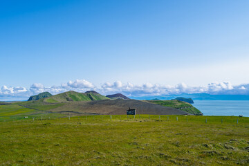Naklejka premium A picturesque view of green grassy hills, a small building, and the ocean under a bright blue sky with fluffy clouds in Vestmannaeyjar, Iceland.