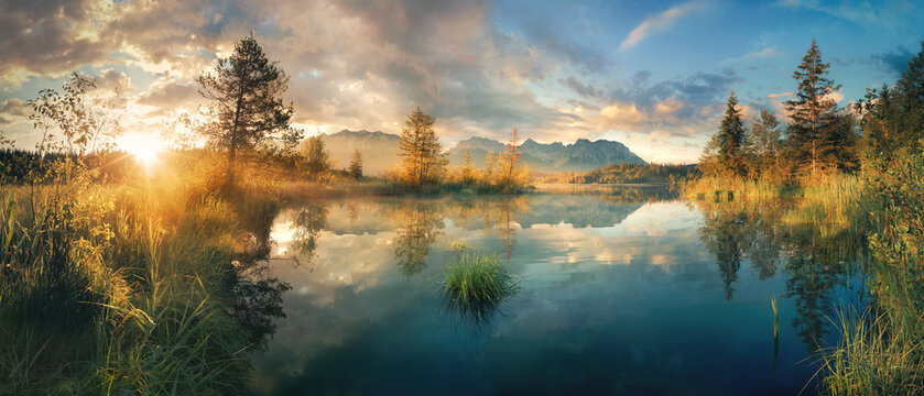 Painterly panoramic lake scenery in Germany at sunrise with mountains, trees, nice colorful sky, beautiful morning sunlight and clear reflections in the calm water 