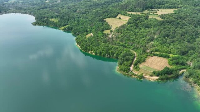 Turquoise water of Butoniga reservoir reflects white clouds in Istria region. Drone flies over artificial lake surrounded by dense green forests and rolling hills near Motovun.