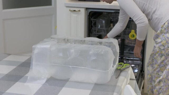 Glass jars sit wrapped on a table while a woman loads a dishwasher in a bright kitchen. She feels calm and focused as she cleans. The neat room brings a sense of peace.