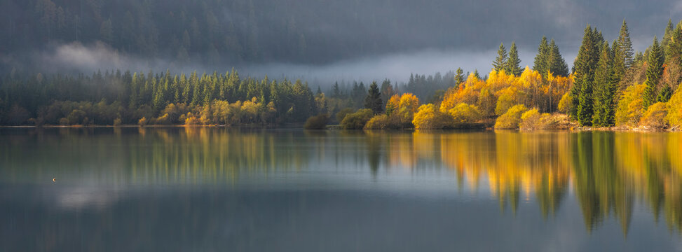 Autumn forest reflecting on tranquil lake surface with mist in nature tourism destination during fall, representing peaceful exploration in the Julian Alps,Predil lake,Tarvisio,Friuli,Italy.