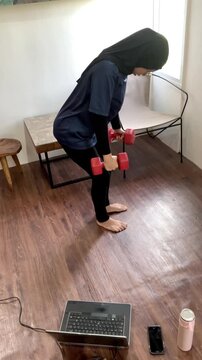 Slow motion vertical video of an Asian Muslim housewife in a hijab doing dumbbell tricep kickbacks. She exercises barefoot at home while watching an online fitness class on her laptop.