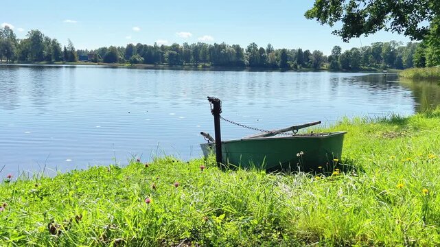 Wide shot of a vintage green rowing boat chained to a post on a grassy lakebank under a bright blue sky on a calm summer day. Countryside nature landscape.