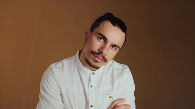 Young adult male with top knot hairstyle and trimmed beard performs series of small pose adjustments while facing camera: starts with hands on hips, leans forward showing forearms, crosses arms
