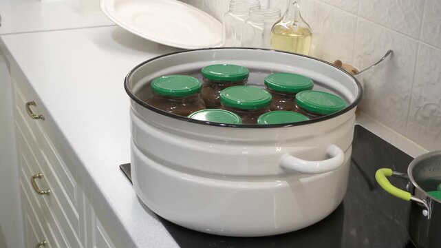 Glass jars with green lids sit inside a large white pot filled with warm water on a stove. This classic canning process fills the bright kitchen with a cozy and peaceful feeling.