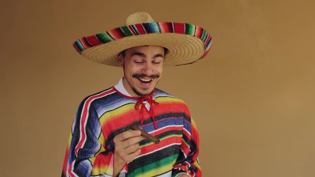 Young man wears woven straw sombrero and bright striped poncho while holding lit cigar near face in studio, with curling smoke, warm light, relaxed expression, cultural festive style