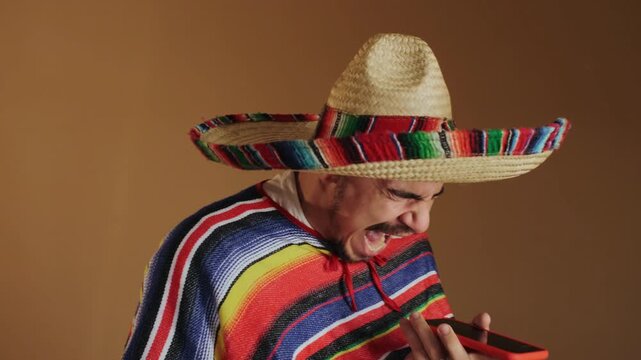 Young adult male wearing woven straw sombrero and bright horizontal striped poncho holds red smartphone at chest level and leans forward while shouting or laughing into device, with man, sombrero