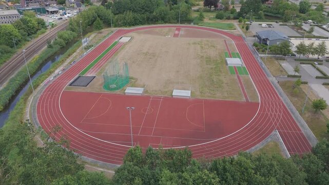 Aerial drone footage circling around an outdoor athletics running track with red lanes, sports field areas, greenery, water and surrounding urban infrastructure. Scenic sports facility viewed from abo