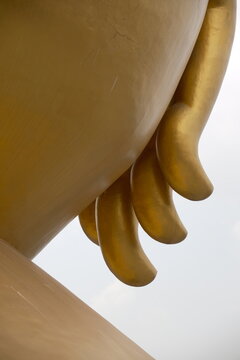 Close up low angle detail of the graceful curved golden fingers and hand of the giant seated Buddha statue resting on the knee against a soft bright white sky at Khao Wong Phrachan in Lopburi Thailand