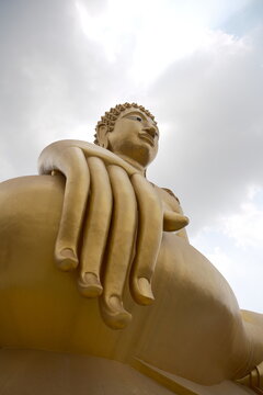 Low angle perspective looking up at the giant golden Buddha statue showing massive fingers resting on the knee and a serene peaceful face against a cloudy sky at Khao Wong Phrachan in Lopburi Thailand