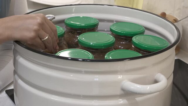 A woman's hand carefully checks glass jars with green lids inside a large white pot. She preserves food in a warm, cozy kitchen setting. The thoughtful task creates a peaceful feeling.