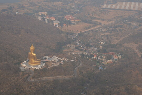 High angle daytime view from the mountain peak at Khao Wong Phrachan in Lopburi Thailand looking down at the giant golden seated Buddha statue and temple complex surrounded by dry forest hills in haze