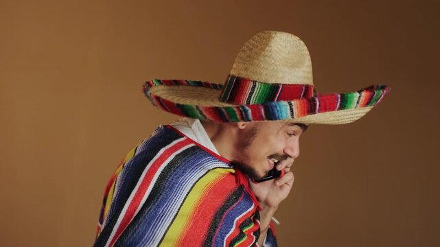 Right-side closeup of young man wearing wide-brim straw sombrero with multicolored trim and striped serape while holding red smartphone to ear and making expressive hand gestures, soft diffused