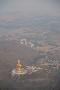 High angle daytime view from the mountain peak at Khao Wong Phrachan in Lopburi Thailand looking down at the giant golden seated Buddha statue and temple complex surrounded by dry forest hills in haze