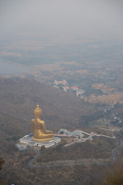 High angle daytime view from the mountain peak at Khao Wong Phrachan in Lopburi Thailand looking down at the giant golden seated Buddha statue and temple complex surrounded by dry forest hills in haze