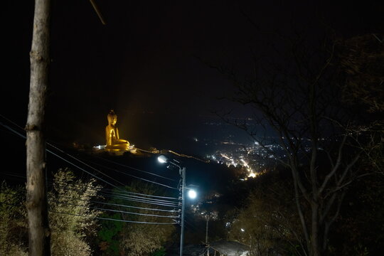 Scenic night view from the mountain trail at Khao Wong Phrachan in Lopburi Thailand showing the giant golden seated Buddha statue overlooking glowing city lights in the valley and a lit stairway below