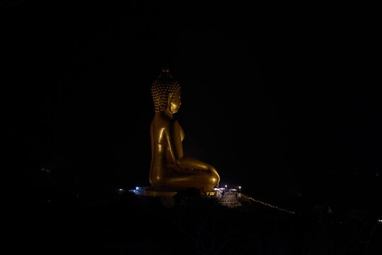 Side profile view of a giant golden seated Buddha statue illuminated at night against the pitch black sky captured from the way up the mountain trail at Khao Wong Phrachan in Lopburi province Thailand
