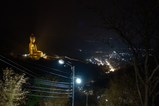 Scenic night view from the mountain trail at Khao Wong Phrachan in Lopburi Thailand showing the giant golden seated Buddha statue overlooking glowing city lights in the valley and a lit stairway below