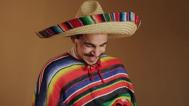 Three-quarter portrait of young man wearing wide woven straw sombrero with multicolor trim and striped serape poncho, seen in studio against warm brown backdrop, with man, sombrero, poncho, phone
