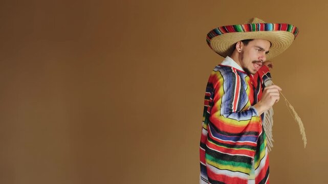 Young man wearing oversized woven straw sombrero with multicolored trim and fringed striped poncho, turning torso and extending right arm forward in presenting or reaching gesture while holding thin