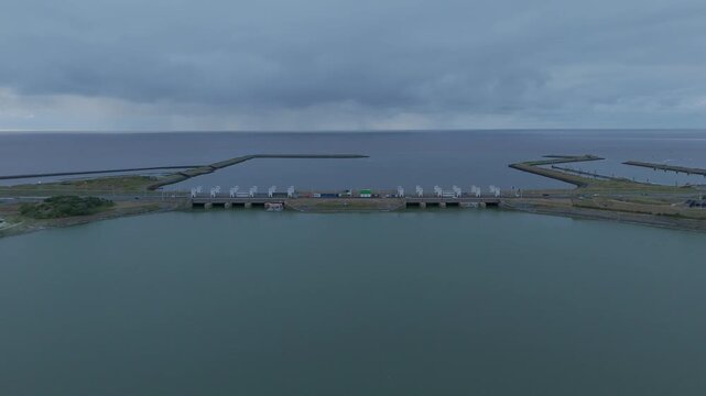 Aerial drone footage flying toward the Lorentz Locks on the Afsluitdijk in the Netherlands, showing road traffic crossing the lock complex between open water under a dramatic cloudy sky.