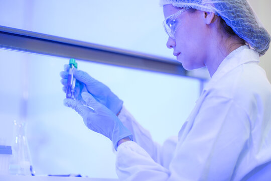 A female research scientist in a white lab coat, hairnet and clear safety goggles bringing the tube closer to her glasses inside a university biotechnology laboratory.