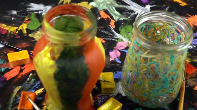 Two empty glass jars painted with acrylic paints, pots with paints, paintbrushes are on a decorative black fabric, home decor, slow motion