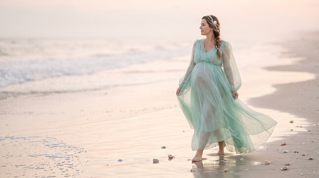 Pregnant woman in flowing mint dress walking on beach at sunset with shells