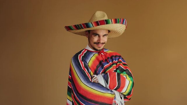 Medium close-up of adult male wearing wide-brimmed woven straw sombrero with multicolor striped band and matching brightly colored serape draped over shoulders, standing against warm brown seamless