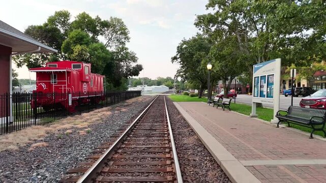 Low forward drone flight gliding down train tracks past a small town depot platform, red caboose, station benches, and a lit lamp post under a soft pale early morning overcast sky.