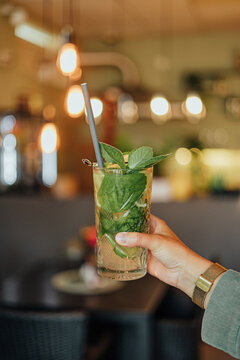Close-up of a hand holding a tall mint and lime drink with straw and garnish against a warmly lit caf&eacute; interior with soft bokeh and copy space