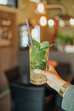 Close-up of a hand holding a tall mint and lime drink with straw and garnish against a warmly lit caf&eacute; interior with soft bokeh and copy space