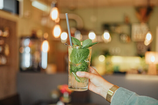 Close-up of a hand holding a tall mint and lime drink with straw and garnish against a warmly lit caf&eacute; interior with soft bokeh and copy space