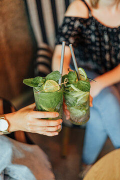 Two people clink glasses filled with green mint and lime drinks while relaxing in caf&eacute; chairs, creating a casual social and refreshing beverage scene