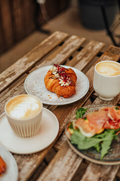 Vertical caf&eacute; scene with decorated croissant, two creamy coffees and prosciutto toast with leafy greens on a weathered wooden table, with copy space