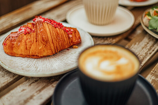 Close-up of a flaky croissant topped with pink icing and red berry pieces beside a creamy coffee on a weathered wooden caf&eacute; table