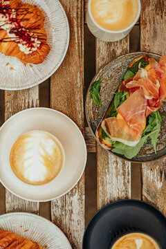 Overhead caf&eacute; table with three coffees, decorated croissants, and an open sandwich topped with arugula and cured ham on rustic wooden slats