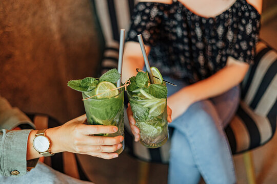 Two people clink glasses filled with green mint and lime drinks while relaxing in caf&eacute; chairs, creating a casual social and refreshing beverage scene