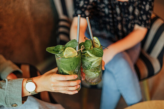 Two people clink glasses filled with green mint and lime drinks while relaxing in caf&eacute; chairs, creating a casual social and refreshing beverage scene