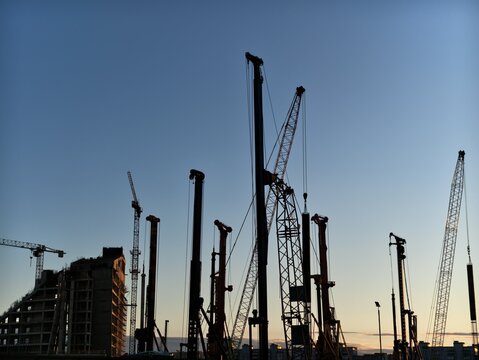 Construction cranes and piling rigs silhouetted against clear evening sky over urban building site, with tall machinery, unfinished structure, and sunset light creating industrial skyline
